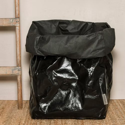 A washable paper bag is shown. The bag is rolled down at the top and features a UASHMAMA logo label on the bottom left corner. The bag pictured is the gigantic size in metallic black. Next to the bag is a wooden ladder.