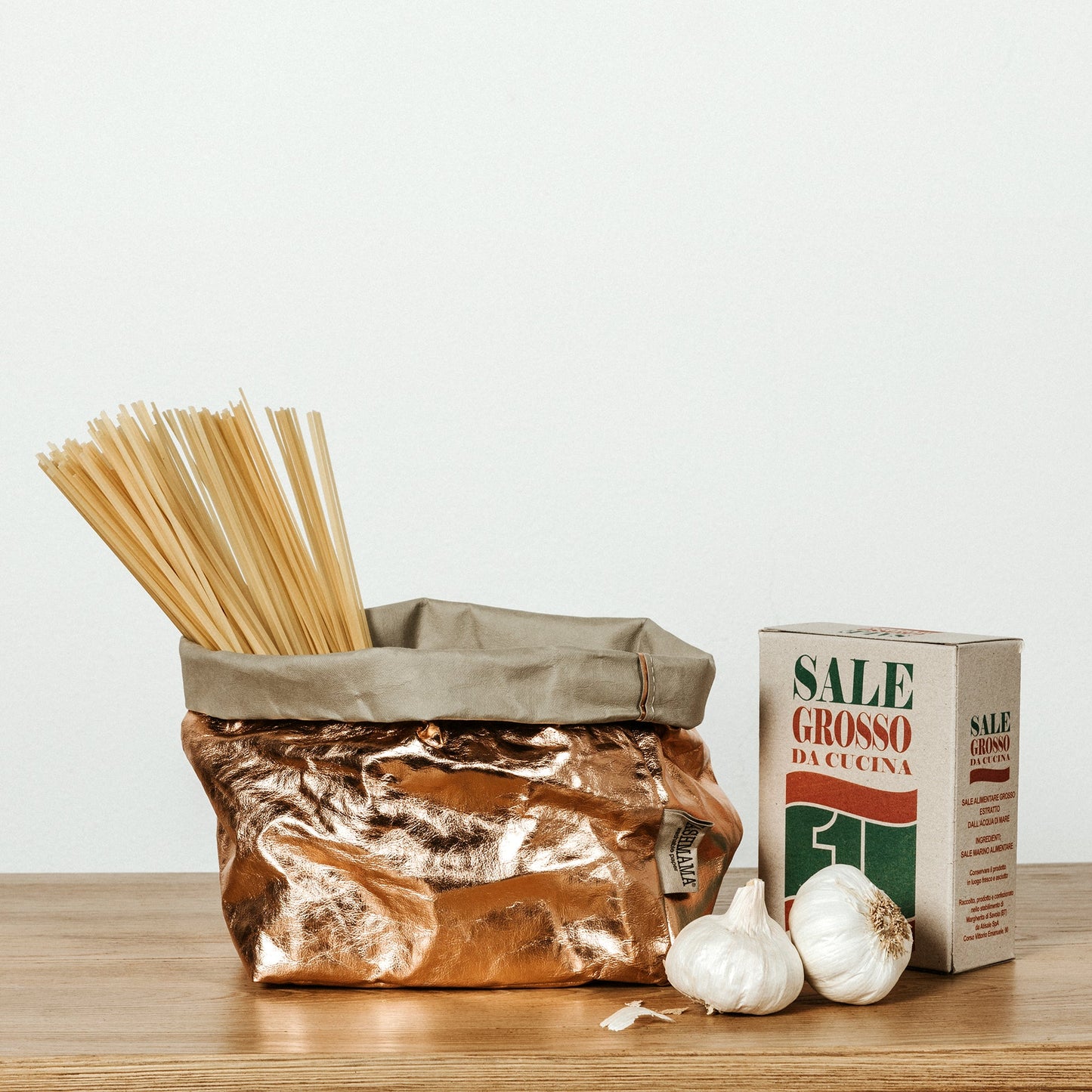 A washable paper bag is shown. The bag is rolled down at the top and features a UASHMAMA logo label on the bottom left corner. The bag pictured is the large size in rose gold metallic. Inside the bag there is some dried spaghetti. Next to the bag there isa box of sale and two bulbs of garlic.