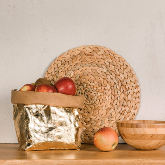 A washable paper bag is shown. The bag is rolled down at the top and features a UASHMAMA logo label on the bottom left corner. The bag pictured is the large plus size in metallic platinum. The bag is shown filled with red apples. Next to the bag are a wooden bowl and an apple. Behind the bag a raffia round placemat is shown leaning against the wall.