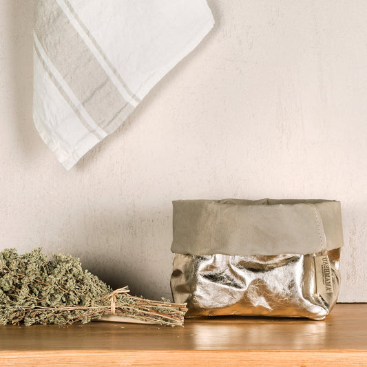 A washable paper bag is shown. The bag is rolled down at the top and features a UASHMAMA logo label on the bottom left corner. The bag pictured is the medium size in metallic platinum. Next to the bag lies a bundle of herbs. On the wall above a linen tea towel is draped.