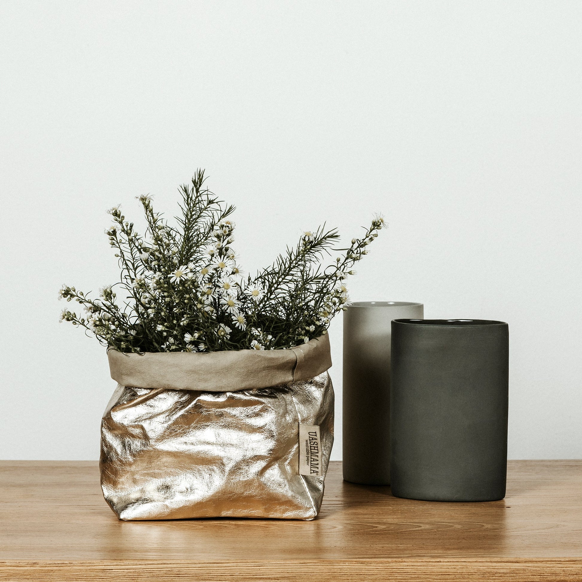 A washable paper bag is shown. The bag is rolled down at the top and features a UASHMAMA logo label on the bottom left corner. The bag pictured is the medium size in metallic platinum. The bag is filled with a flowering plant. Next to the bag are two pillar candles in grey and black.