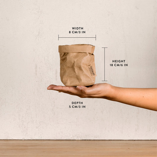 A washable paper bag is shown. The bag is rolled down at the top and features a UASHMAMA logo label on the bottom left corner. The bag pictured is the piccolo size in tan. The dimensions are shown on the image. The bag is shown being held in the palm of a woman's hand.