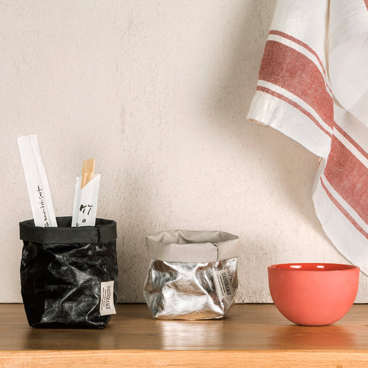 Two piccolo washable paper bags are shown. The one on the left is metallic black and contains two sets of chopsticks. The one on the right is metallic silver and is shown empty. To the right is a small pink bowl. Draped in the background is a linen tea towel.