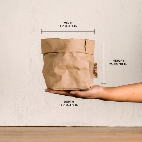A washable paper bag is shown. The bag is rolled down at the top and features a UASHMAMA logo label on the bottom left corner. The bag pictured is the small size in tan. The dimensions are shown on the image. The bag is shown being held in the palm of a woman's hand.
