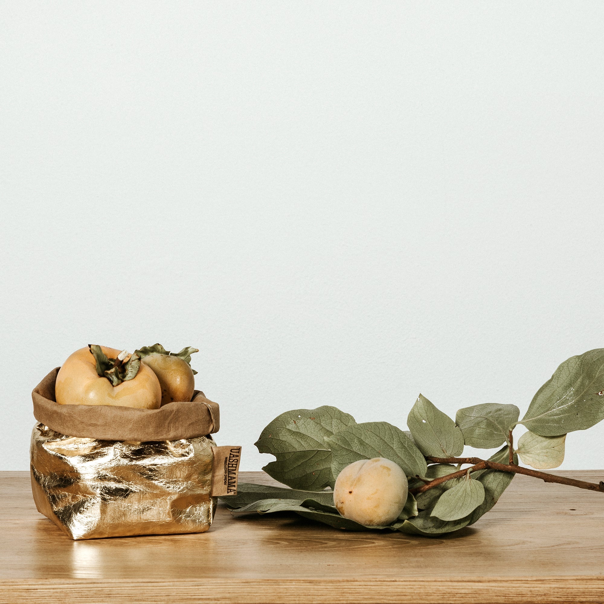 A small metallic gold washable paper bag is shown filled with persimmons. Next to the the bag is an olive branch and a piece of fruit.