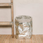 A washable paper bag is shown. The bag is rolled down at the top and features a UASHMAMA logo label on the bottom left corner. The bag pictured is the extra large size in metallic platinum. Next to the paper bag is a wooden ladder.