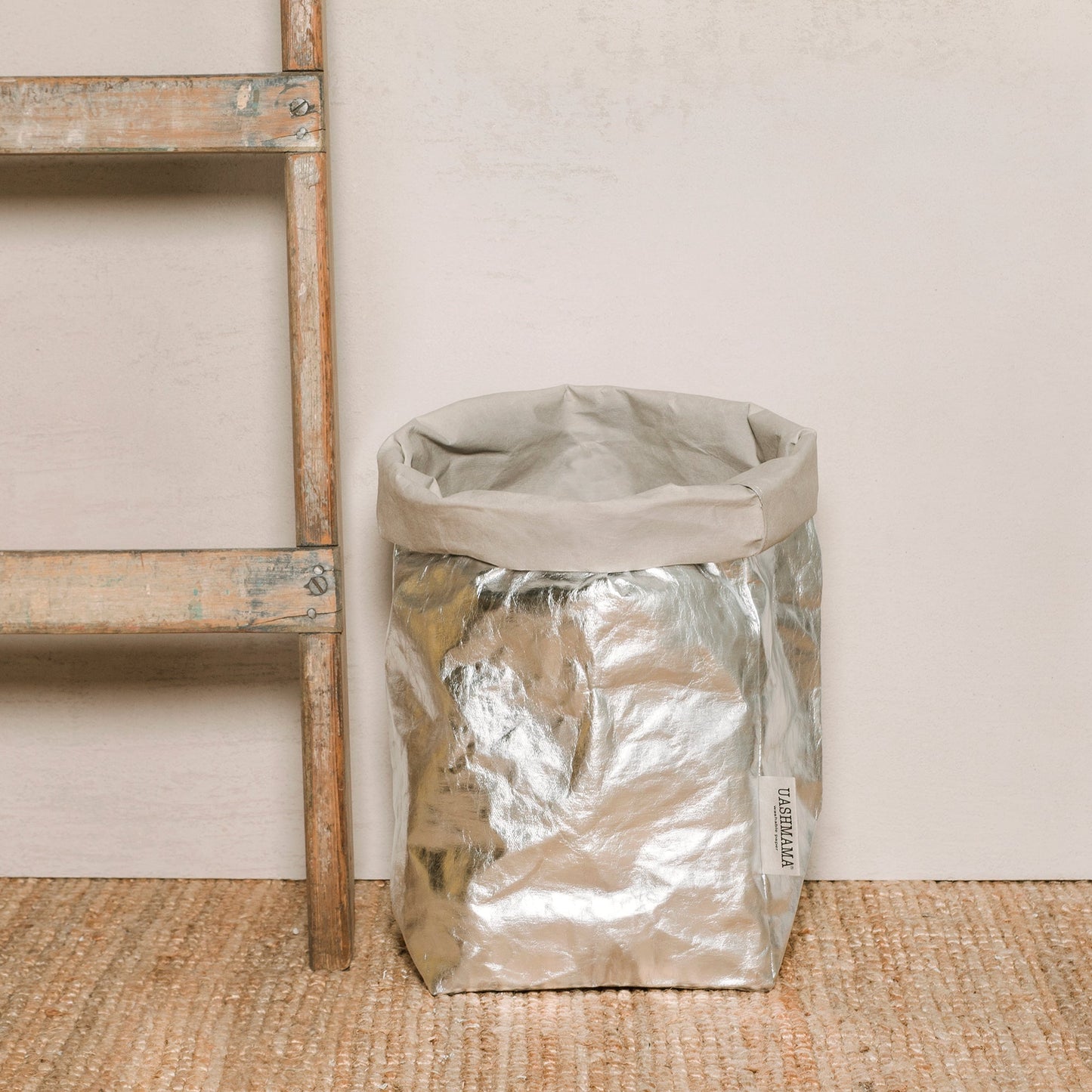 A washable paper bag is shown. The bag is rolled down at the top and features a UASHMAMA logo label on the bottom left corner. The bag pictured is the extra large size in metallic platinum. Next to the paper bag is a wooden ladder.