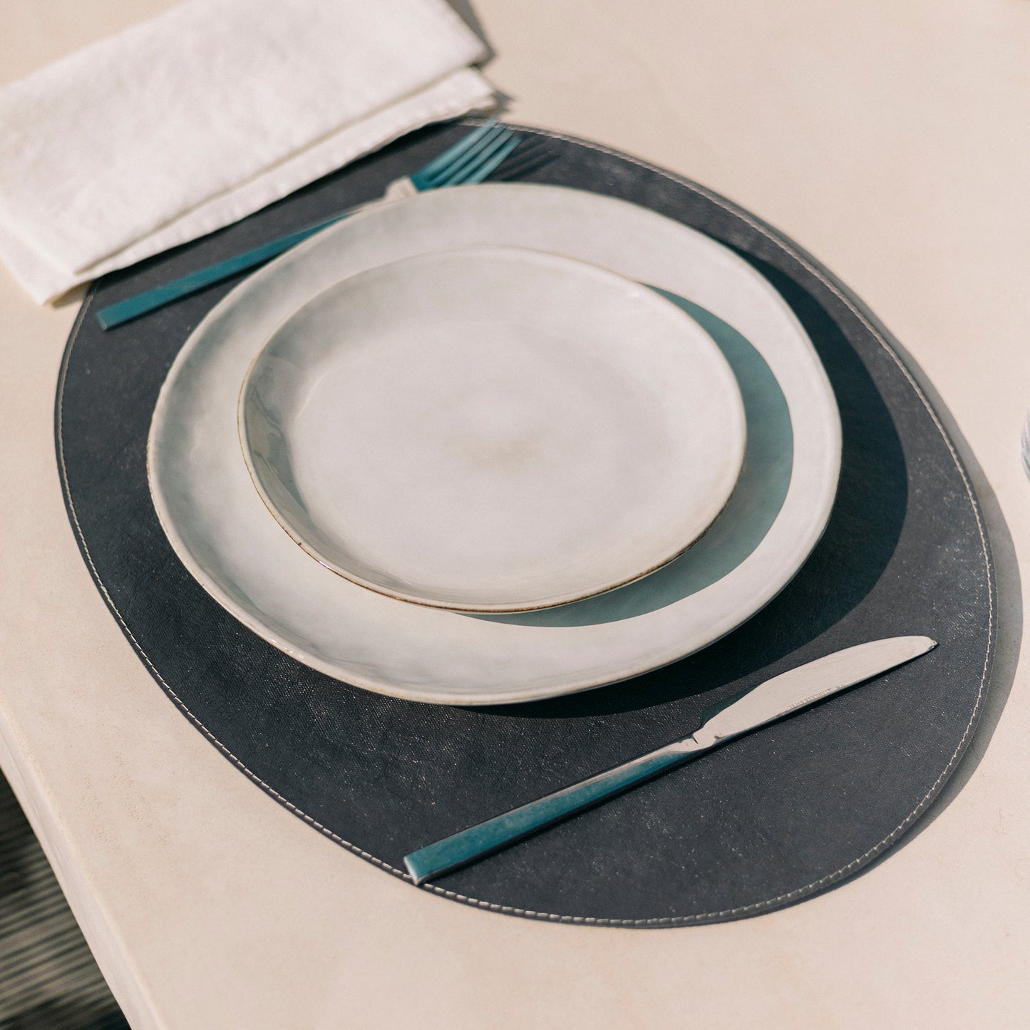 A place setting is shown on a white outdoor dining table. A dark grey oval washable paper placemat is shown with two white stoneware plates stacked on top. A fork and knife are shown beside the plates, and a white linen napkin is also resting on the placemat.