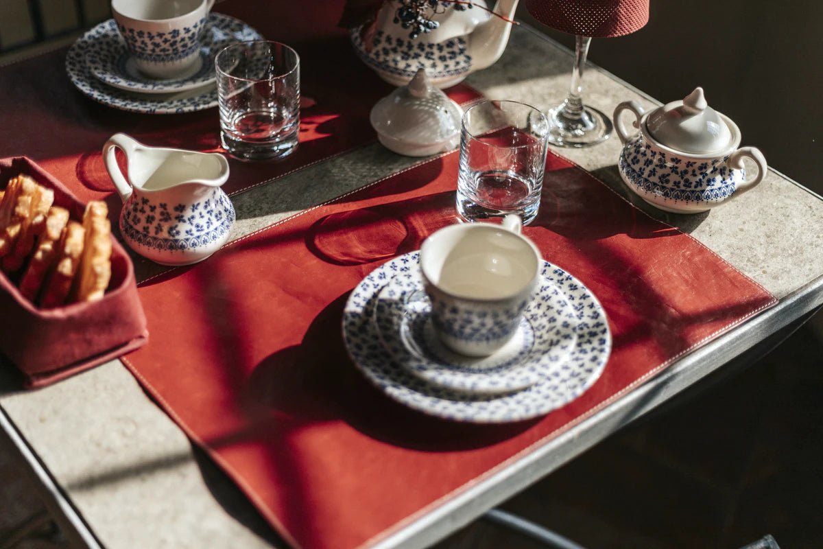 A breakfast table is set for two. Two red washable paper rectangular placemats are shown with a plate and cup and saucer stacked on each one. In there same white and blue china pattern, there are also a tea pot, sugar pot and milk jug shown on the table, as well as two drinking glasses. On the left hand side of the table there is a red washable paper tray containing slices of a sweet bread.