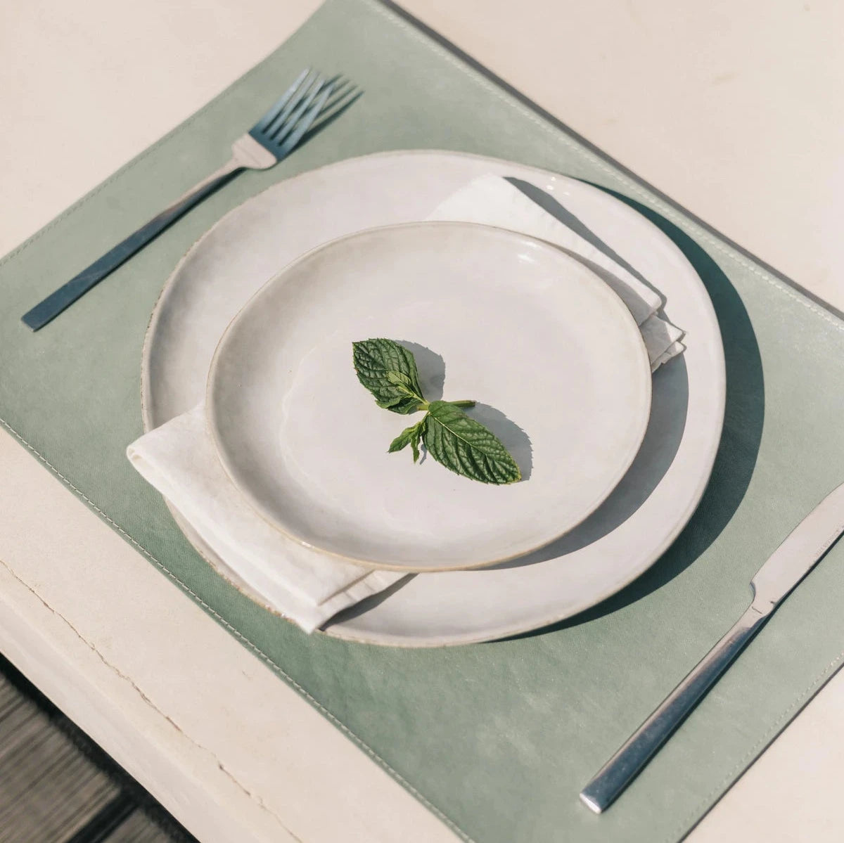 The image shows a white outdoor dining table and one place setting. The place setting consists of a pale green washable paper rectangular placemat, a large stoneware plate, a linen napkin and a smaller stoneware plate on top. Resting on the smaller plate are two basil leaves. On the left of the plate s a silver fork and a silver knife lies to the right.