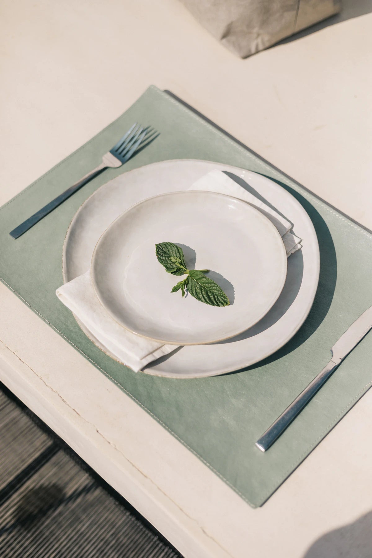 The image shows a white outdoor dining table and one place setting. The place setting consists of a pale green washable paper rectangular placemat, a large stoneware plate, a linen napkin and a smaller stoneware plate on top. Resting on the smaller plate are two basil leaves. On the left of the plate s a silver fork and a silver knife lies to the right.