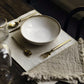 The image shows a dark wooden dining table with one place setting. The place setting is a cream washable paper rectangular place mat, set with a cream stoneware bowl and set of gold cutlery. Next to the fork is a linen napkin and in the top right corner of the placemat is a drinking glass.