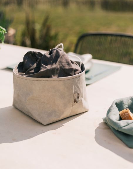 An outdoor dining table is shown. On the table is a washable paper pale grey bread bag with a dark grey linen drawstring top. Behind the bag is a pale green washable paper placemat, and next to the bag is an organic linen green bread bag with a slice of bread inside.