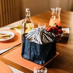 A wooden dining table is shown set with orange washable paper square placemats. In the middle of the table on one of the placemats is a blue washable paper bread bag with a striped blue and white cotton drawstring top. The bag is shown closed. Also on the table is a glass bottle of water, a jar containing fruit juice and a plate.