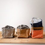 Four washable paper square bread bags with drawstring tops are shown from the side. On the left is a pale grey washable paper bread bag, then a tan coloured washable paper bread bag, then next is an orange washable paper bread bag. Stacked on top of the orange bag is a blue washable paper bread bag. All the bags are shown closed with the drawstrings cinched.