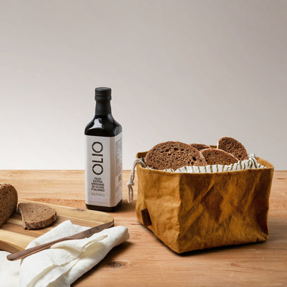 A tan washable paper square bread bag with a striped cotton drawstring top is shown open and filled with small slices of bread. Next to the washable paper bag is a bottle of olive oil, a wooden chopping board with some slices of bread, a silver knife and a linen napkin.