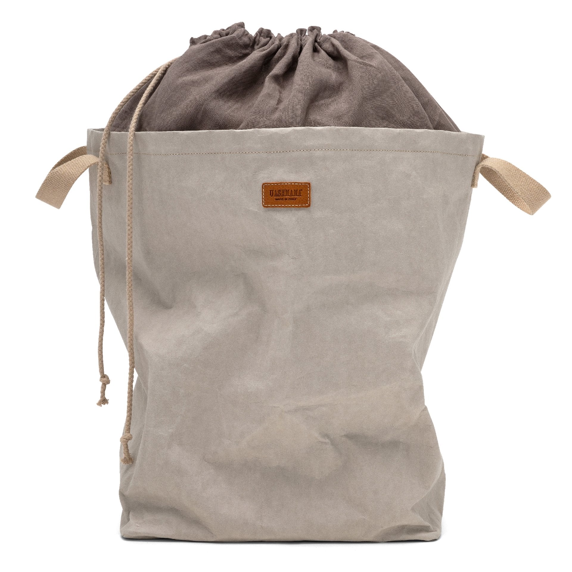 A washable paper laundry bag with a drawstring linen top is shown. The bag has two small recycled cotton handles on either side at the top of the bag, and one drawstring closure at the top. A small leather UASHMAMA logo leather label is on the front of the bag. The bag is pale grey in colour with a dark grey linen top.