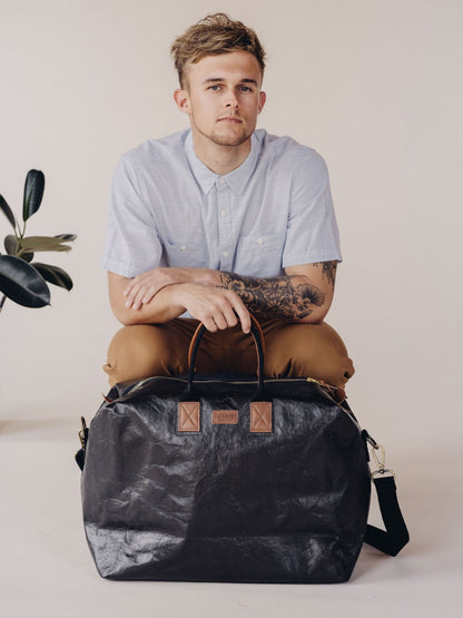 A man is shown sitting on a low stool. In front of him on the floor is a large washable paper holdall in black with tan leather details.