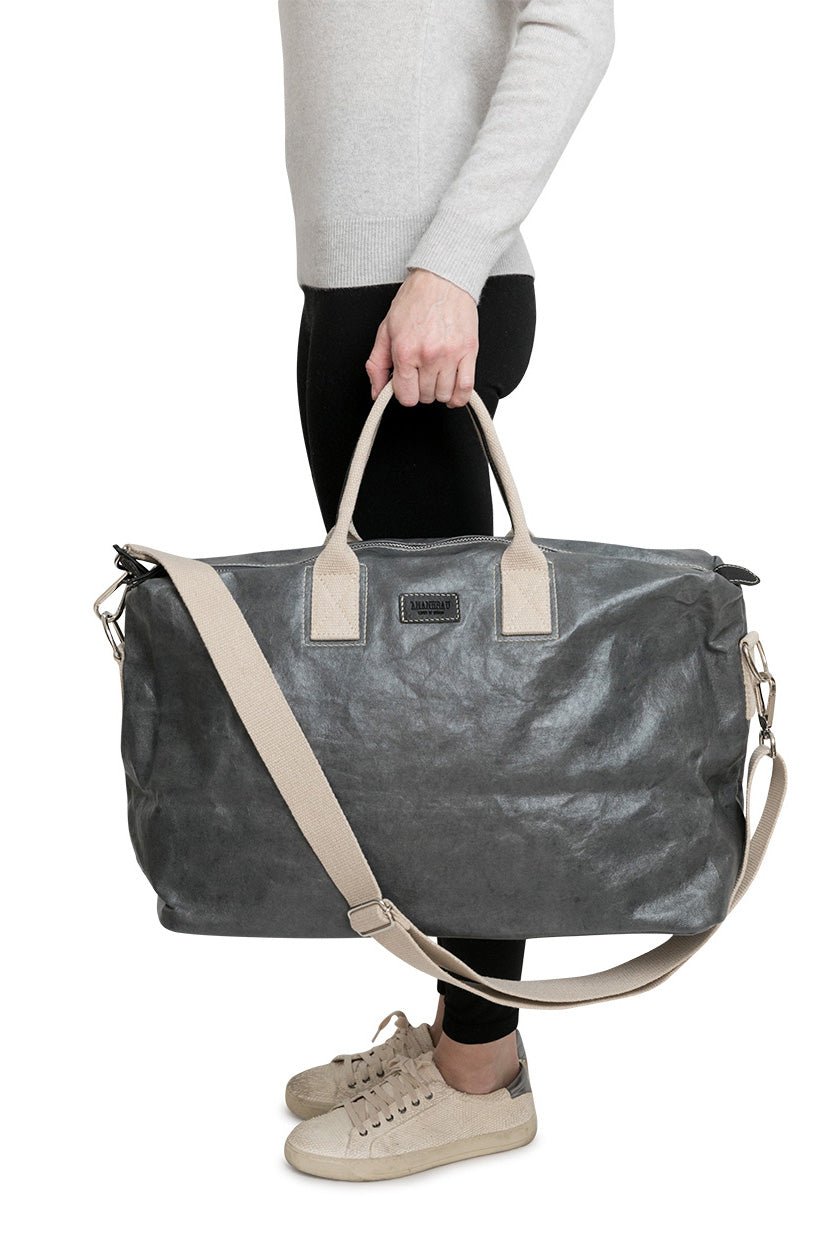 A person is standing facing sideways. In their left hand they are carrying a dark grey large washable holdall with cream recycled cotton handles and a recycled cotton shoulder strap.