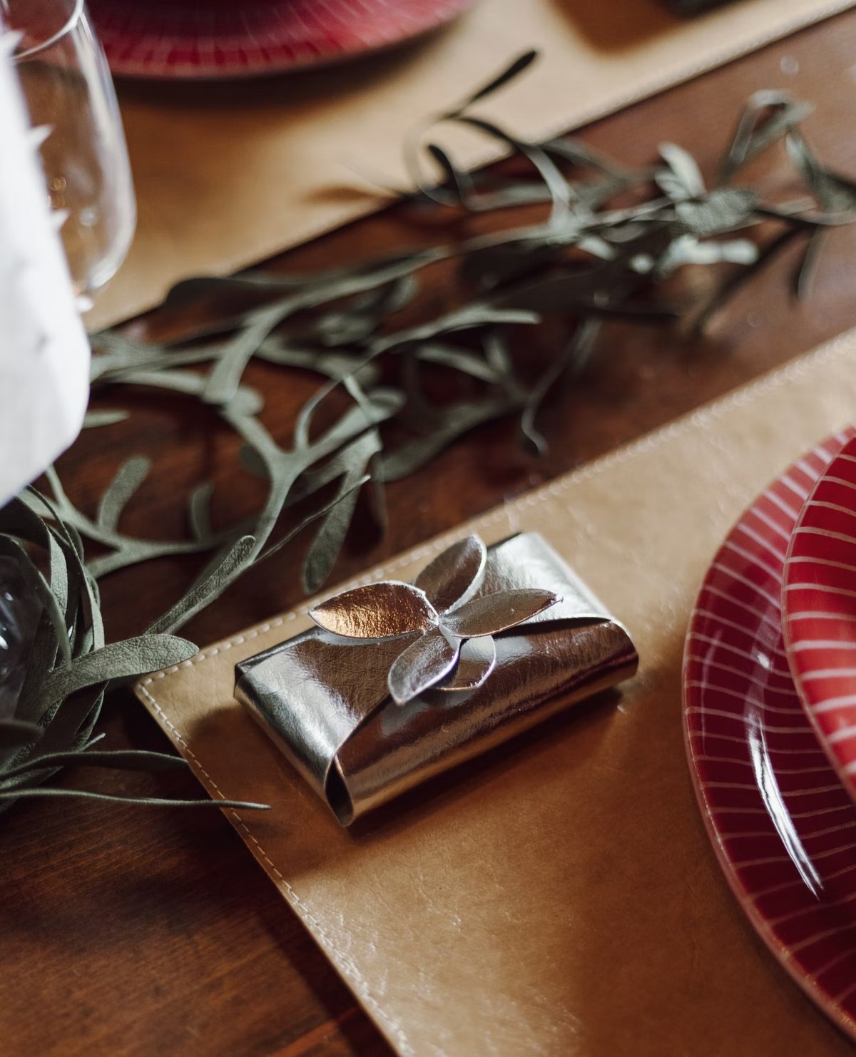 A wooden dining table is shown in close up. The table is set with tan washable paper placemats and red dishes. On one of the placemats is a metallic dark grey soap holder, closed with a petal fastening.