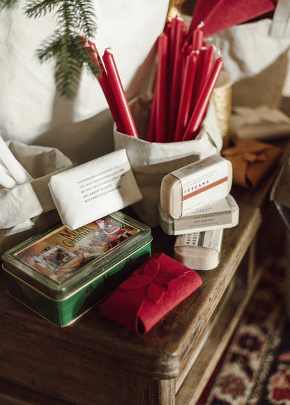 A wooden table is shown decorated for Christmas. On the table is a cream washable paper bag containing red taper candles. Also shown are three bars of soap stacked up and a white and red washable paper soap packaging. The packaging is shown closed with a folded petal closure.