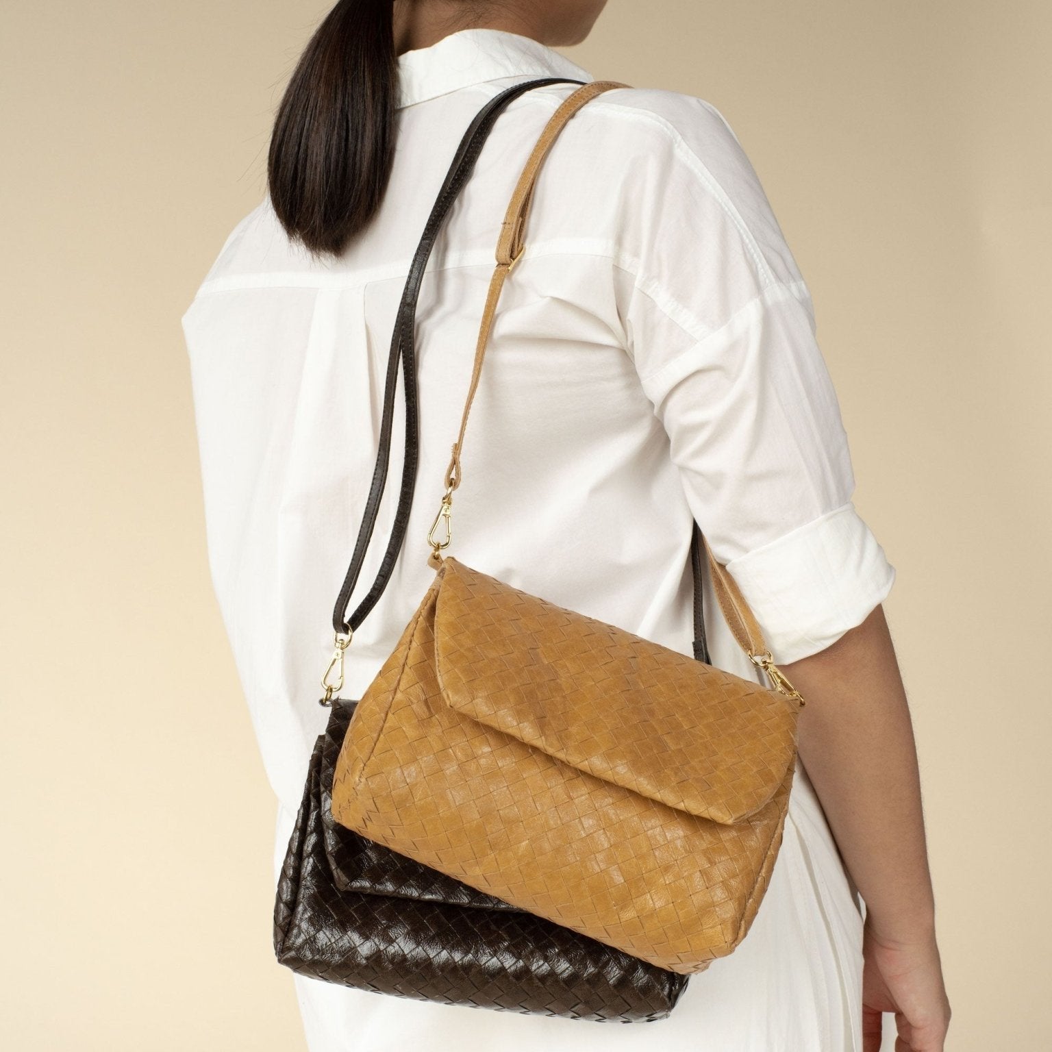 A woman is shown with her back to the camera. Over her right shoulder she is carrying two woven washable paper handbags, with washable paper shoulder straps and a front flap closure. The bags are Black and Tan in colour.