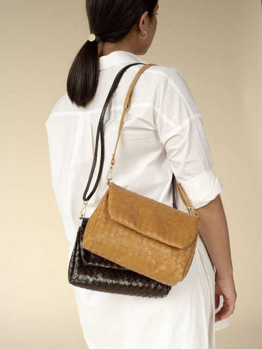 A woman is shown with her back to the camera. Over her right shoulder she is carrying two woven washable paper handbags, with washable paper shoulder straps and a front flap closure. The bags are Black and Tan in colour.