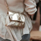 The torso of a woman is shown. She is wearing jeans and a cream sweater, and she is facing away from the camera. Across her body she is wearing a small washable paper handbag with a front flap closure. The bag is metallic platinum in colour.