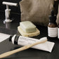A sink is shown with some beauty products next to it. A brown washable paper toiletry bag is shown along with a bottle of facial serum, a bar of soap on a soap dish and a tube of toothpaste with a bamboo toothbrush leaning on the tube.