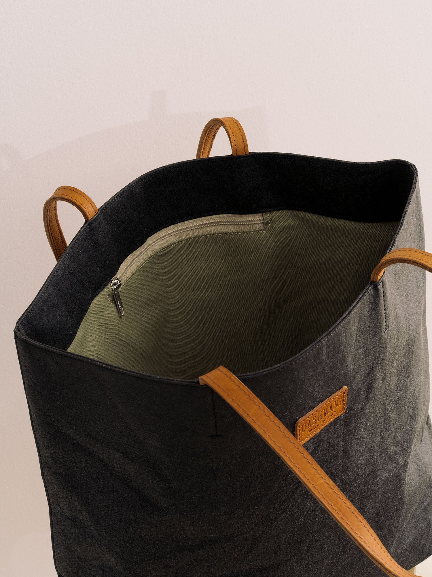 A black washable paper tote bag with tan handles and a leather UASHMAMA logo label is shown open. The image shows an organic cotton lining and an internal zipped pocket.