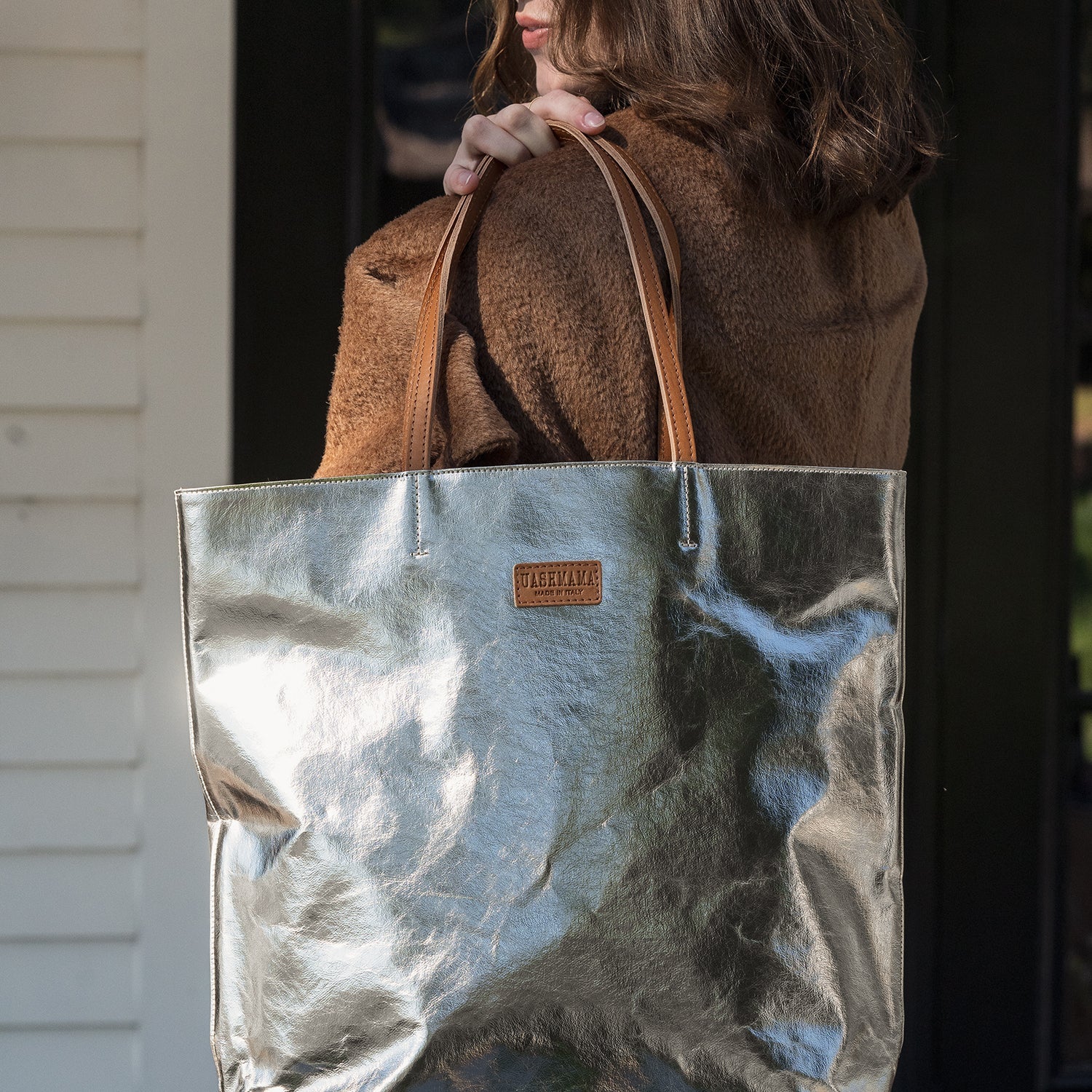 A woman is shown standing looking backwards over her shoulder. She is holding a platinum metallic washable paper tote bag in her right hand, draped over her left shoulder. The bag has two tan leather handles and a tan leather UASHMAMA logo label. 
