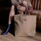 A woman is shown sitting on a brown leather bench seat. She is holding a brown washable paper tote bag by its two tan leather handles. The bag has an open top and a small tan leather UASHMAMA logo label on the front.