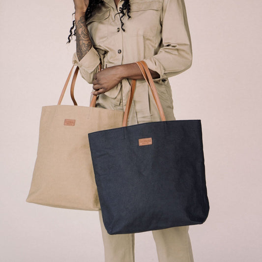 A woman is shown standing holding two washable paper tote bags, one in black and one in tan. The tote bags have leather handles, an open top and a small UASHMAMA leather logo label on the front.