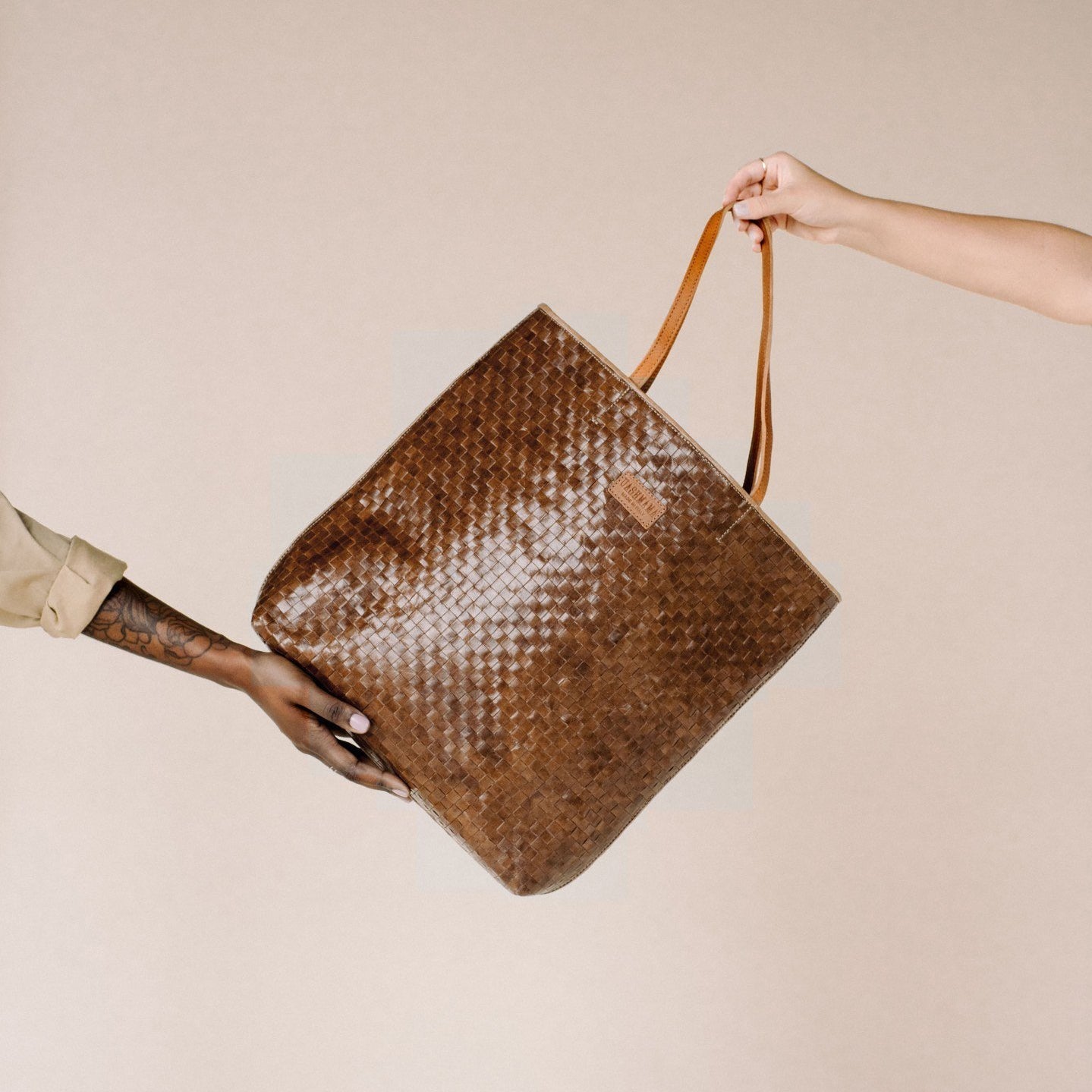 A dark tan woven washable paper tote bag with tan leather handles and a tan leather UASHMAMA logo label, is shown being passed from one woman's hand to another. One hand is flat along the base of the bag, and the other hand is shown holding the handles.