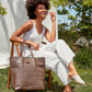 A woman is sitting in a wooden chair outside in a garden. She is smiling and wearing summer clothes. On her right forearm she is carrying a dark brown woven washable paper tote bag with brown leather handles and small UASHMAMA leather logo label.
