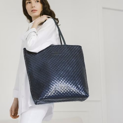 A woman is standing and wearing white. She is carrying a dark blue woven paper tote bag over her left shoulder. The bag has dark blue leather straps and a dark blue UASHMAMA leather logo label.