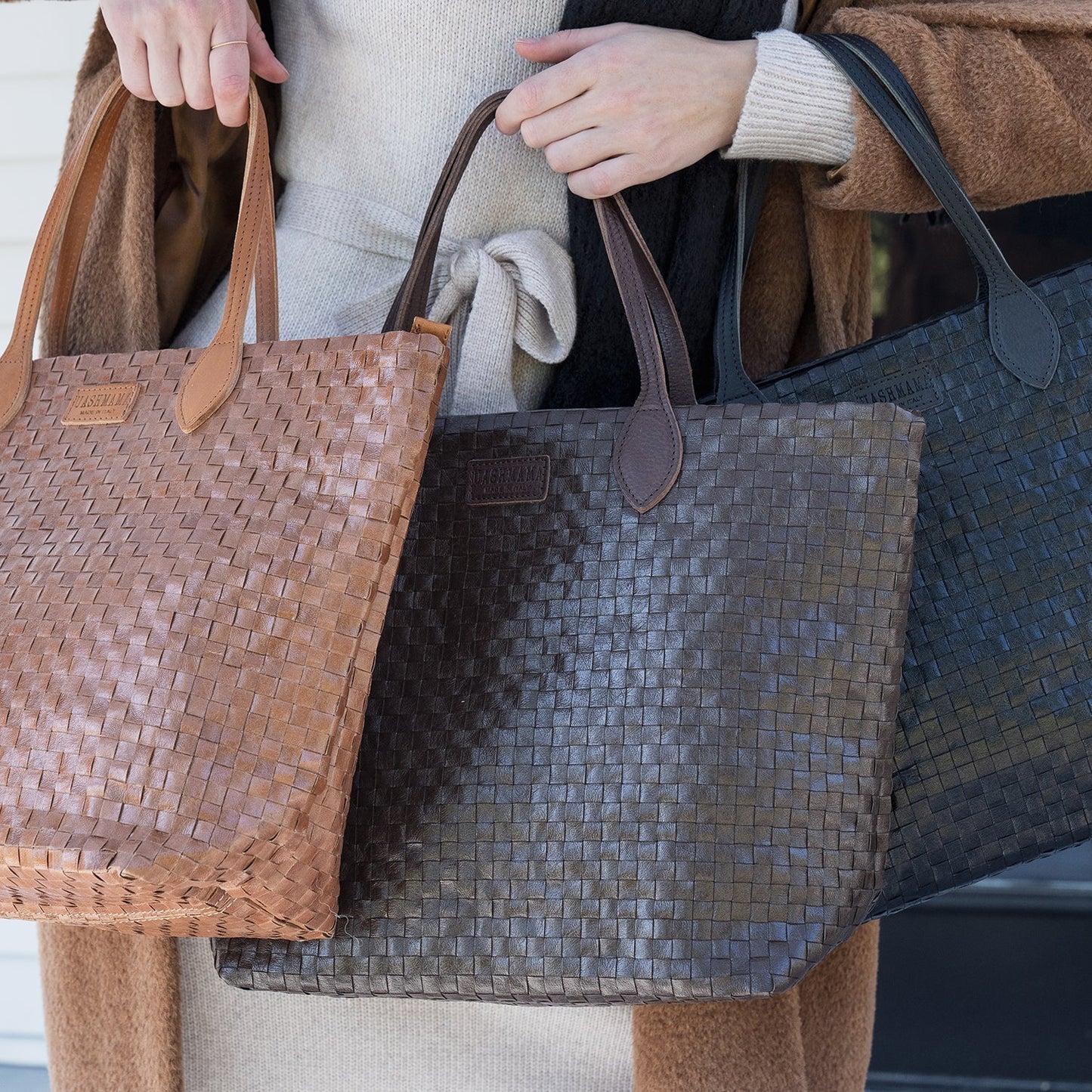 A woman is shown carrying three large washable paper woven tote bags. The bags are black, dark tan and brown in colour and all have long leather handles and a UASHMAMA leather label in between the handles. The tote bags close with a zip.