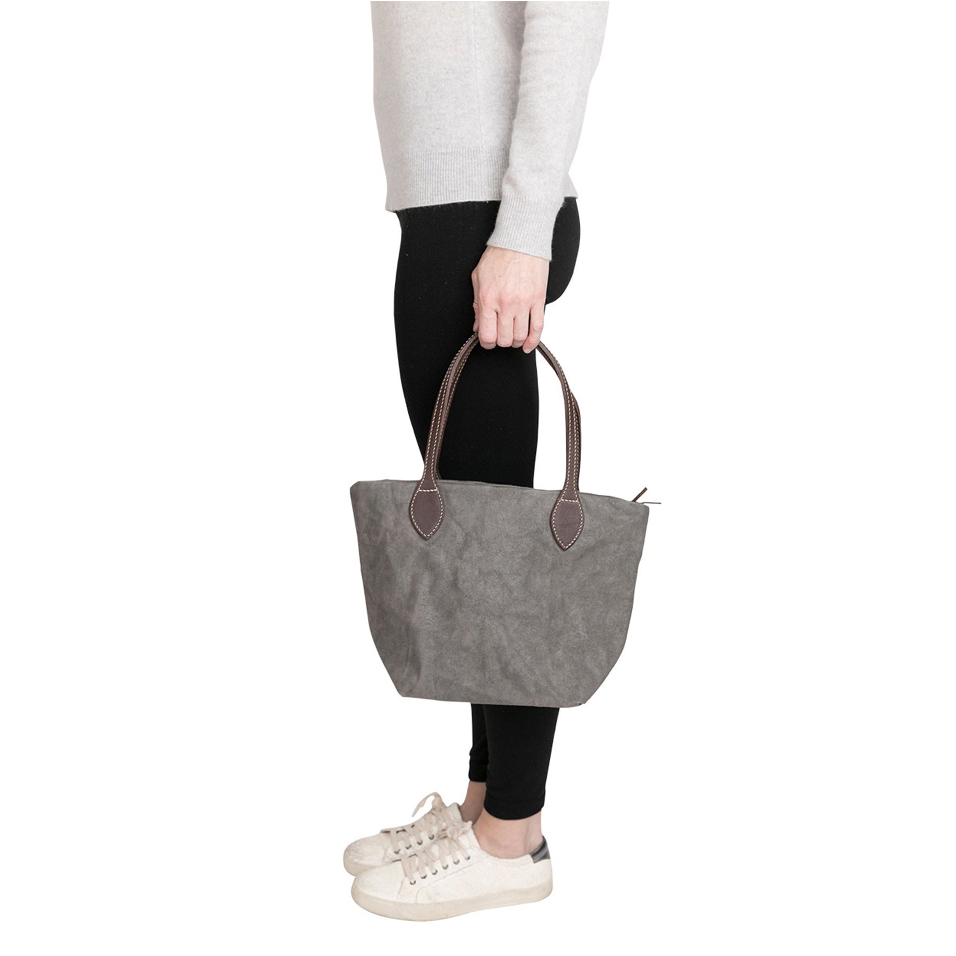 A woman is shown standing facing sideways. She is carrying a small washable paper tote bag. It has two long dark grey leather handles. The bag shown is dark grey.