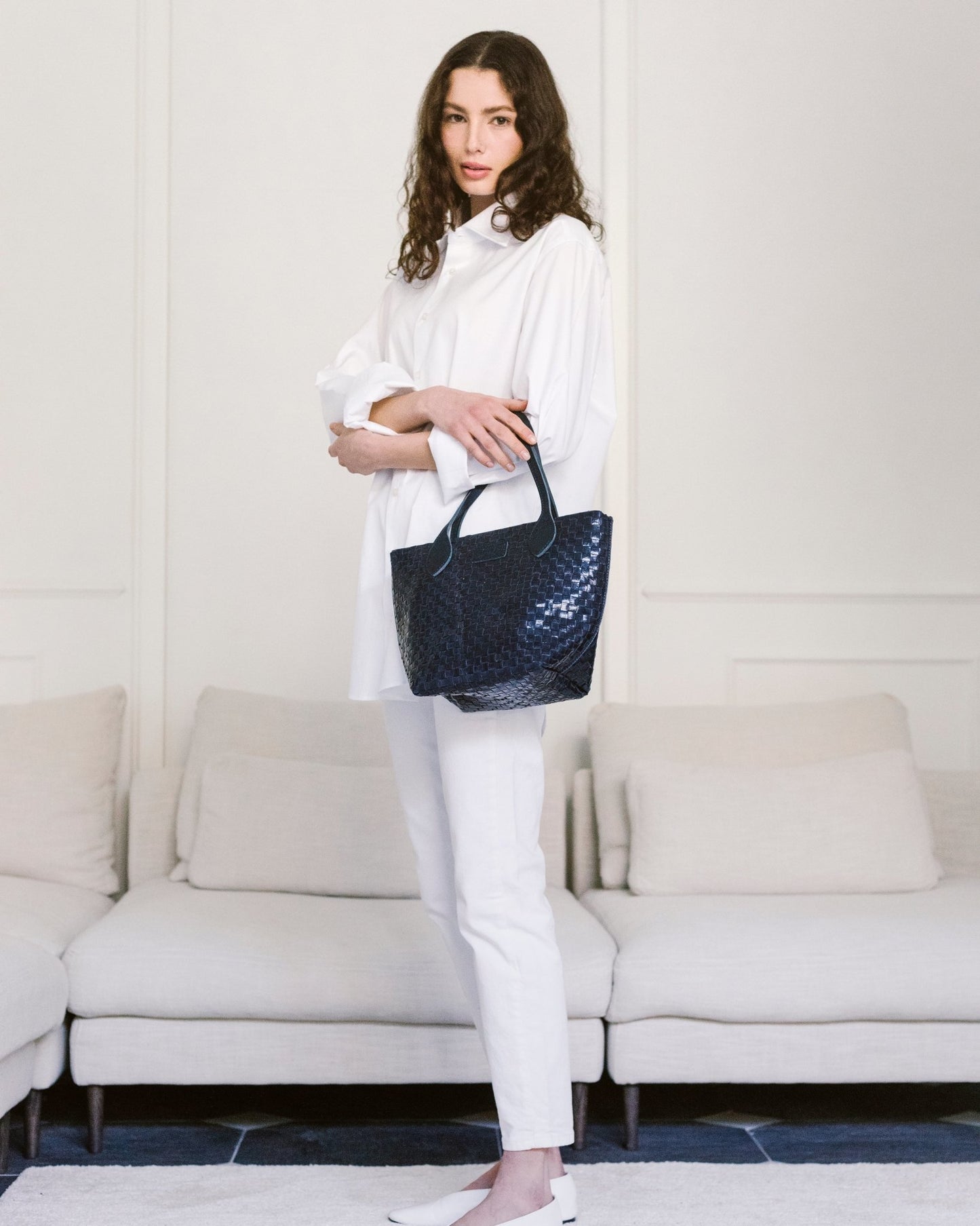 A woman is standing in front of a white sofa. She is wearing a small UASHMAMA washable paper woven tote bag on her left arm. The bag is dark blue.