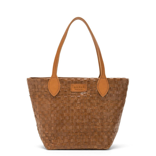 A small washable paper woven tote is shown. It has two long tan leather handles and a tan leather UASHMAMA logo label on the outside in between the handles. The bag shown is in a dark tan colour.