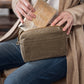 A woman in a cardigan and jeans is shown sitting. Across one shoulder she is wearing an olive coloured washable paper handbag with an external pocket. She is shown sliding a metallic washable paper wallet into the open handbag.