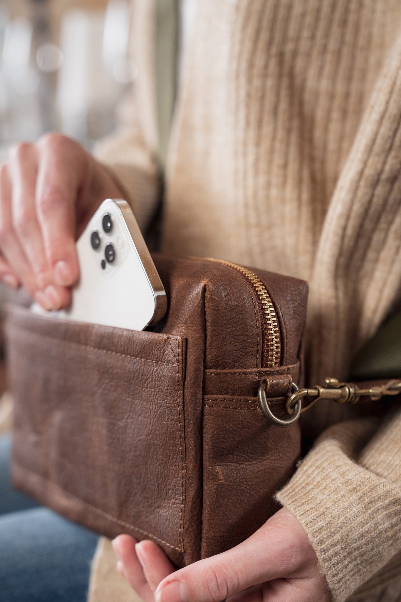 A woman is shown sitting holding a brown washable paper cross body bag with an external pocket. She is sliding an iPhone into the external pocket of the bag.