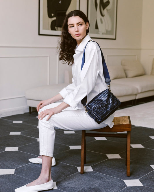 A woman is shown sitting on a wooden stool with her hands on her lap. On her shoulder is a woven washable paper square shaped handbag. The bag is dark blue in colour with a wide dark blue fabric shoulder strap.