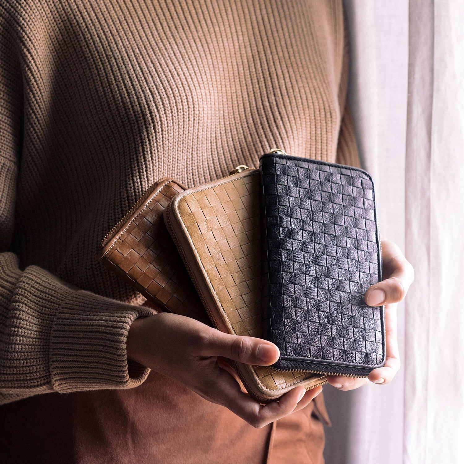 A woman is shown standing. In her hands she is holding three large zipped washable paper woven wallets. The wallets are dark tan, light camel and black in colour.