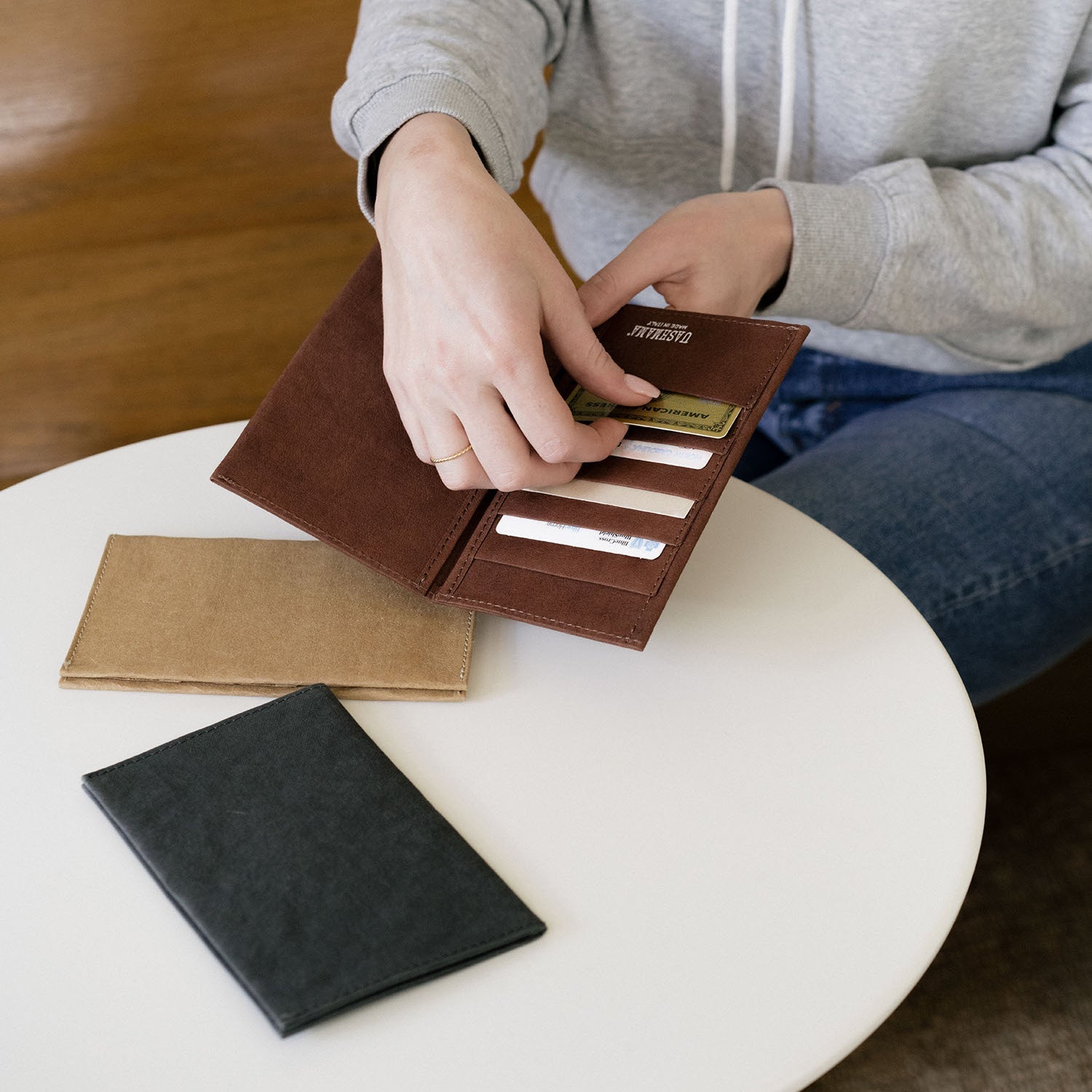 A woman in a grey sweatshirt and jeans is shown sitting at a round white table. On the table are two washable paper large wallets in tan and black. The woman is holding a washable paper wallet in dark brown in her hand and is inserting a credit card into one of the 5 card slots.