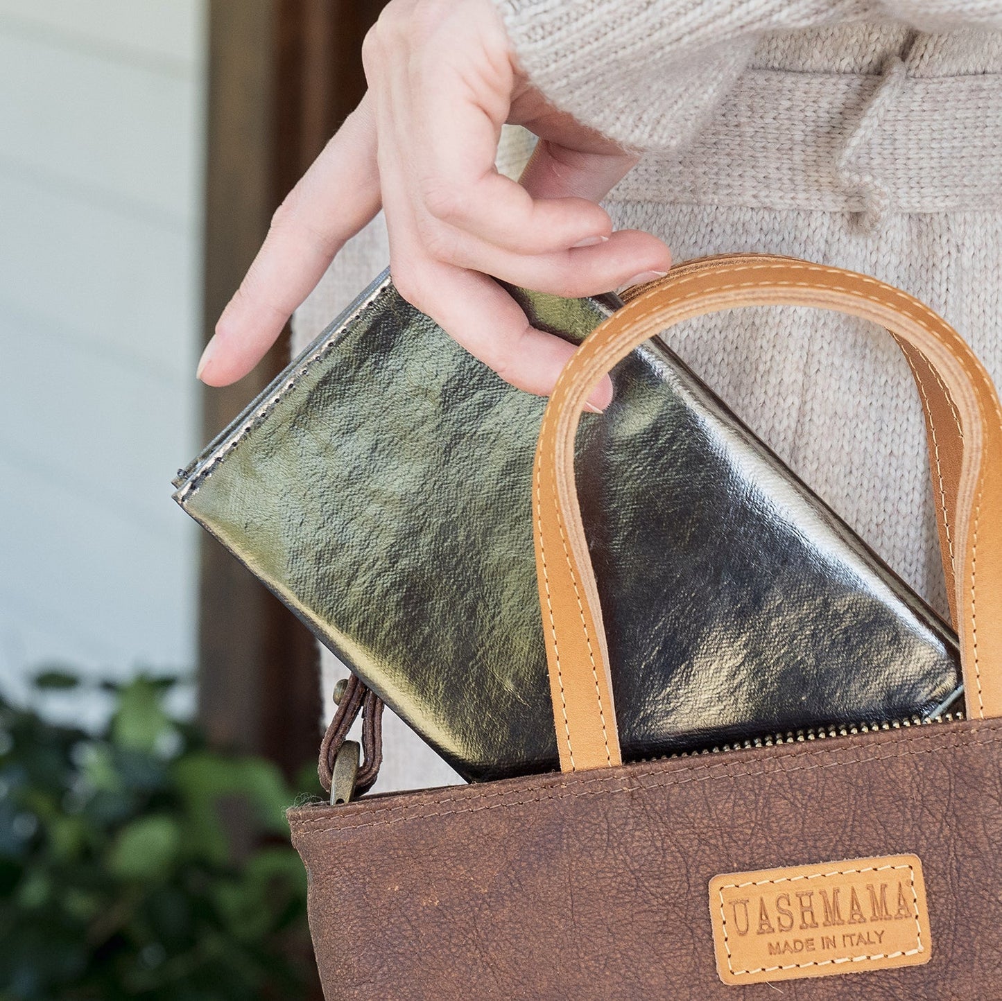 A woman is shown inserting a dark metallic grey large washable paper wallet, into a dark brown washable paper handbag which has the UASHMAMA logo in a tan leather tag on the front.