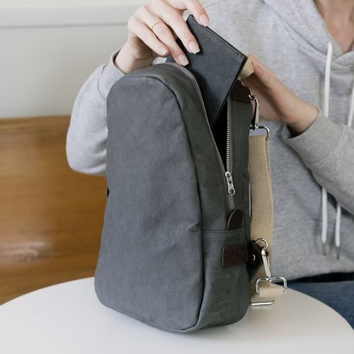 A woman in a grey sweatshirt is shown. On the table next to her is a dark grey washable paper backpack. She is inserting a small dark grey washable paper wallet into the backpack.