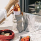 A woman is shown lifting a bottle of rose wine out of a pale grey washable paper bag. Also shown on there table are a silver wine cooler with the UASHMAMA logo, a washable paper tray containing strawberries, and a red linen bread bag containing bread rolls.