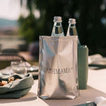 A table is set with washable paper placemats. On the table is a green linen bread bag, a green washable paper ice cube bottle cooler, and a silver UASHMAMA bottle cooler, containing a bottle of water.