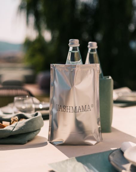 A table is set with washable paper placemats. On the table is a green linen bread bag, a green washable paper ice cube bottle cooler, and a silver UASHMAMA bottle cooler, containing a bottle of water.